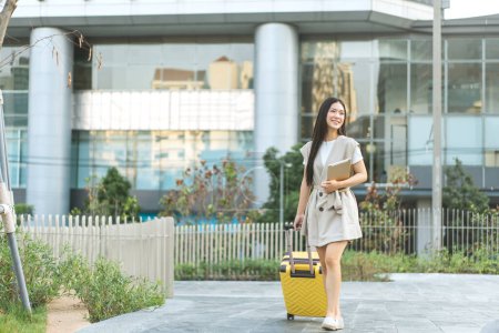 Foto de Mujer asiática adulta joven independiente caminando con equipaje de viaje y tableta digital para viaje de negocios. Hermosa sonrisa cara pelo largo. Los estilos de vida urbanos la gente en el día. Fondo de la ciudad - Imagen libre de derechos