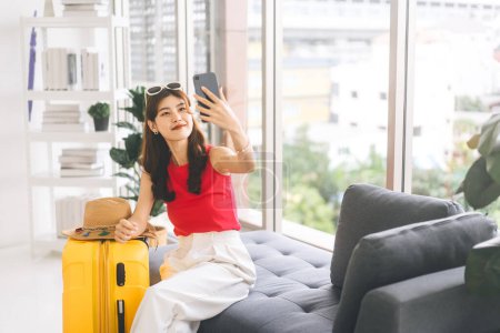 Téléchargez les photos : Portrait de jeune femme adulte du sud-est asiatique avec des bagages de voyage jaunes. Utilisation du smartphone pour selfie avant de commencer le voyage. Se préparer pour le voyage à la maison concept. - en image libre de droit