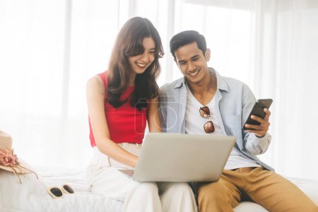Photo for Focus on woman. Young adult southeast asian lover couple using laptop for getting ready for holidays travel trip. Living at home or hotel sitting on bed with hat and luggage. Happy holidays journey. - Royalty Free Image