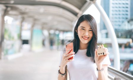 Foto de Portrait of smile young adult business working asian woman. Hand holding mobile phone and house model for online insurance application concept. Background with copy space on day. - Imagen libre de derechos