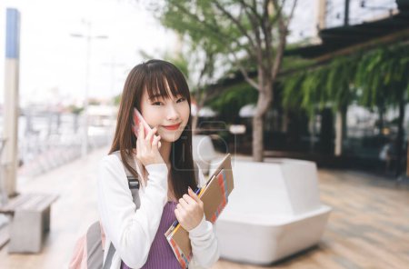 Téléchargez les photos : Collège gens style de vie sur le concept de jour. Portrait de jeune étudiante asiatique heureuse utilisant un téléphone portable. Tenir la papeterie portable et porter un sac à dos à l'extérieur. - en image libre de droit