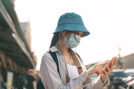 Téléchargez les photos : Jeune adulte asiatique femme sac à dos voyageur porter chapeau et masque facial pour covid-19. Femme utilisant téléphone portable applicaiotn. Les personnes voyageant en ville style de vie à l'extérieur après obtenir le vaccin. - en image libre de droit