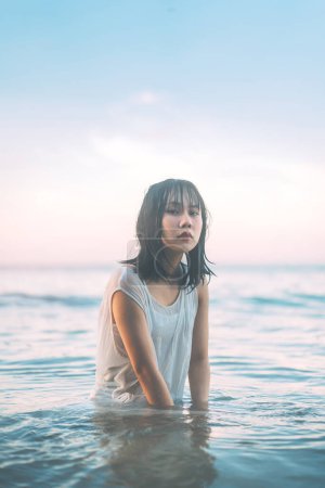 Téléchargez les photos : Portrait de femme asiatique à la plage avec les cheveux mouillés. Assis dans les vagues de la mer Fond de lumière du soleil. Style de vie en plein air sur le concept jour. - en image libre de droit