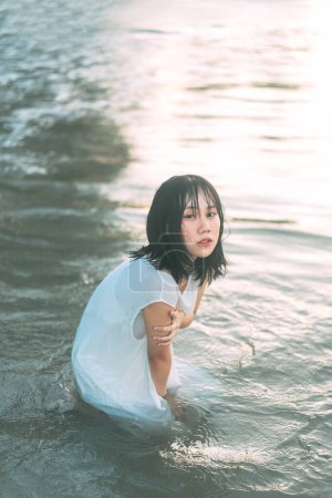 Téléchargez les photos : Portrait de femme asiatique à la plage avec les cheveux mouillés. Assis dans les vagues de la mer Fond de lumière du soleil. Style de vie en plein air sur le concept jour. - en image libre de droit