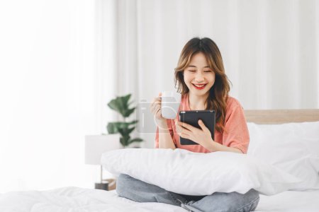 Photo for People lifestyle in bedroom after wake up in morning concept. Young adult single asian woman with coffee using digital tablet for social message telemedicine and mental health or shopping online. - Royalty Free Image