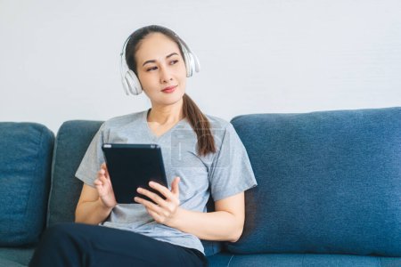 Photo for People happy lifestyle satisfation at home concept. Young adult asian woman wear headphone listen music and relax using digital tablet. Sitting on blue sofa. - Royalty Free Image