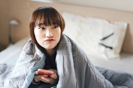 Téléchargez les photos : Portrait de jeune adolescente asiatique. Réveillez-vous dans la chambre à coucher et en utilisant un téléphone mobile. Elle a les cheveux longs et la couverture de couverture grise - en image libre de droit