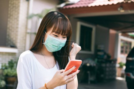 Téléchargez les photos : Adolescente asiatique portant un masque facial pour un nouveau mode de vie normal. Fille à l'aide d'un smartphone et heureux virus corona ou covid 19 vaccin est le succès. Extérieur sur fond de jour. - en image libre de droit