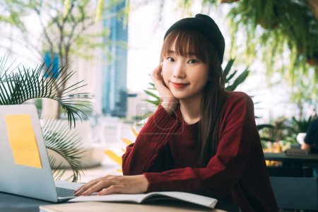 Photo for Portrait young adult asian college student woman wear red sweater and wool hat. Using laptop study and work online at cafe. University people city lifestyle at outside from home on winter day. - Royalty Free Image