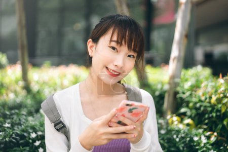 Foto de College people lifestyle on day time concept. Young adult asian student woman using mobile phone for online application at outdoor. - Imagen libre de derechos