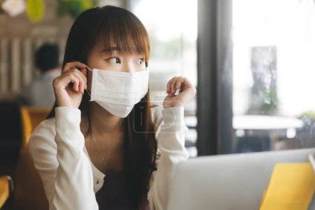 Foto de Young adult asian female with protective face mask for virus corona or covid 19. Work and study at cafe new normal during pandemic lifestyle concept. Indoor background on day - Imagen libre de derechos