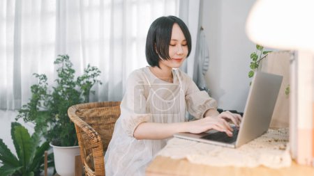 Photo for People lifestyles at living home apartment concept. Young asian woman using laptop computer relax with digital technology. Cozy room style with plant wood table and lamp light. Girl with casual cloth - Royalty Free Image