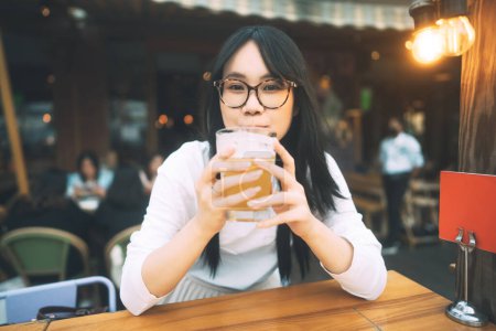 Foto de Retrato de mujer joven asiática adulta con anteojos bebiendo cerveza en el restaurante al aire libre. La gente se relaja en el concepto de descanso. - Imagen libre de derechos