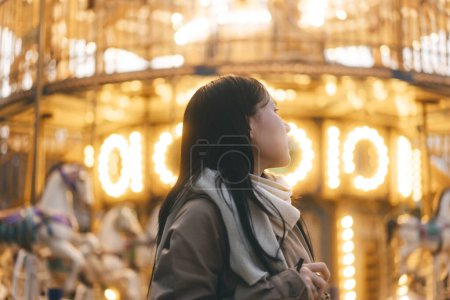 Foto de Retrato de mujer asiática al aire libre en invierno en la ciudad con carrusel o alegre ir alrededor de fondo. Noche de iluminación con luz de neón. La gente se relaja en el concepto de descanso. - Imagen libre de derechos