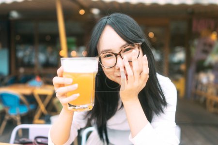 Photo for Portrait of young adult asian woman with eyeglasses drinking beer at restaurant outdoor area. People relax in city break concept. - Royalty Free Image