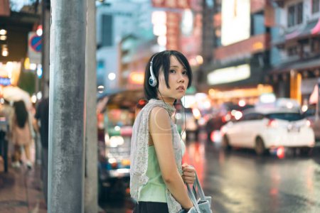 Photo for Urban real people lifestyles in downtown concept. Young asian woman wear y2k nostalgia trend outfit stylish with headphone. Standing at asia chinatown street food night market Bangkok, Thailand - Royalty Free Image