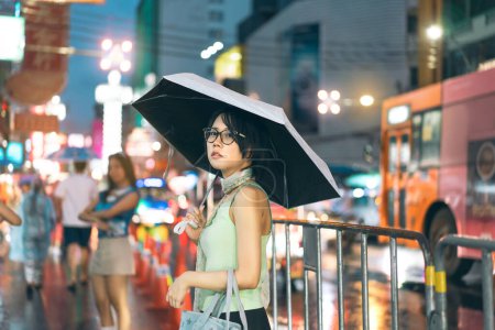 Téléchargez les photos : Urban real people lifestyles in downtown concept. Jeune femme asiatique porter y2k tenue tendance nostalgie élégant. Utiliser le parapluie pendant la saison des pluies. Debout à Chinatown marché de nuit Bangkok ville Thaïlande - en image libre de droit