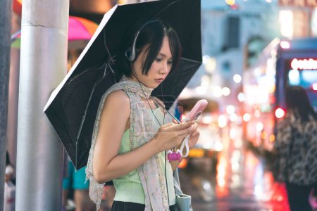 Photo for Urban real people lifestyles in downtown concept. Young asian woman wear y2k nostalgia trend outfit stylish using headphone and flip phone. Rainy season with umbrella. Stand in Bangkok city Thailand - Royalty Free Image