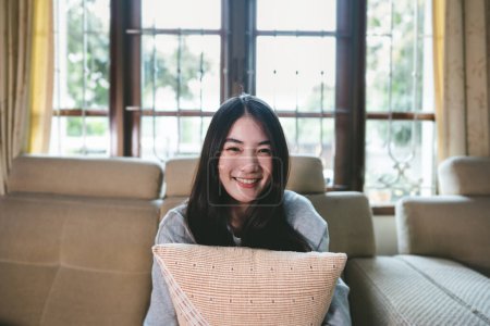 Foto de Retrato de feliz joven adulto mujer asiática cara sonriendo con los dientes. Mirando la cámara sentada en el sofá en casa. Ventana de desenfoque luz natural blackground. - Imagen libre de derechos