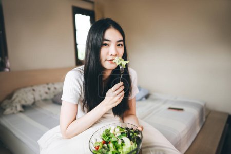Foto de Feliz joven mujer asiática adulta comer desayuno ensalada de verduras en el dormitorio en la mañana. Dieta saludable estilo de vida con concepto de felicidad. Fondo con espacio de copia. - Imagen libre de derechos