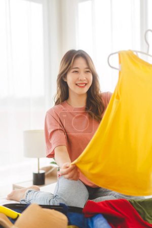 Photo for Vertical portrait of young adult southeast asian dyed hair woman traveler with yellow shirt. Packing a backpack for travel trip. Sitting on bed at home or hotel bedroom. - Royalty Free Image