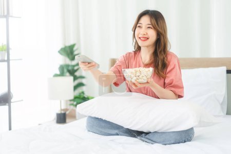 Photo for Joy of a young Asian woman watching a movie in the bedroom. With a smile on her face, she uses the remote to control the TV while enjoying popcorn. A moment of relaxation and entertainment. - Royalty Free Image
