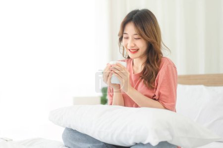 Téléchargez les photos : Sérénité d'une jeune femme asiatique tenant une tasse de café. Profitez de l'heure paisible du matin alors qu'elle est assise sur un lit blanc, avec un sourire heureux. Moment de détente et de contentement. - en image libre de droit