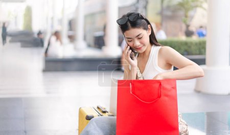 Foto de La gente de la ciudad estilos de vida con la compra de consumismo de compras. Joven mujer asiática adulta hablando con smartphone. Cara de sonrisa feliz sentado en los grandes almacenes. Relájese en vacaciones. - Imagen libre de derechos