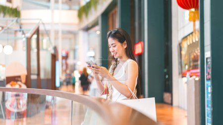Photo for People city lifestyles with buying shopping consumerism. Young adult asian woman using smartphone typing message. Happy smile face sitting at indoors department store. - Royalty Free Image