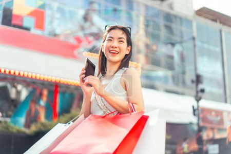 Foto de La gente de la ciudad estilos de vida con la compra de consumismo de compras. Joven mujer asiática adulta usando teléfono inteligente para la banca billetera digital. Cara sonrisa feliz de pie al aire libre. - Imagen libre de derechos