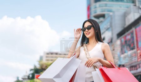 Foto de La gente de la ciudad estilos de vida con la compra de consumismo. Joven cara hermosa mujer asiática bolsas de compras usan gafas de sol. Cara sonrisa feliz de pie en grandes almacenes al aire libre. - Imagen libre de derechos