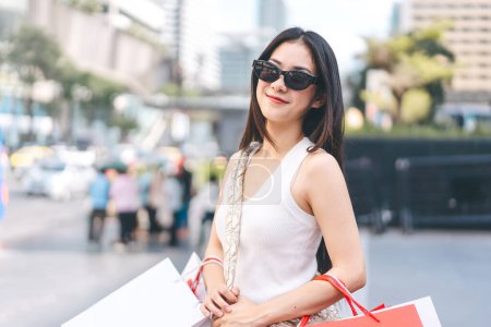 Foto de La gente de la ciudad estilos de vida con la compra de consumismo. Joven cara hermosa mujer asiática bolsas de compras usan gafas de sol. Cara sonrisa feliz de pie en grandes almacenes al aire libre. - Imagen libre de derechos