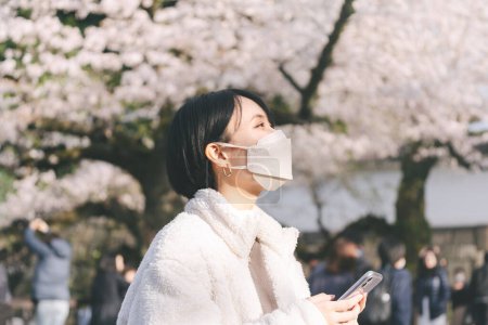 Foto de Joven adulto asiático japonés mujer usar mascarilla usando smartphone mirando hacia arriba en sakura árbol. Viajar en Japón en primavera. - Imagen libre de derechos