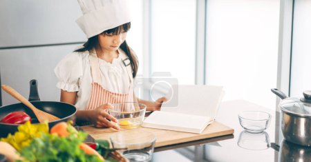 Téléchargez les photos : Petite fille asiatique avec chapeau de chef et tablier cuisine avec recette. Loisirs modes de vie avec des aliments faits maison à la maison. Croissance des enfants avec intérêt concept de compétence. - en image libre de droit