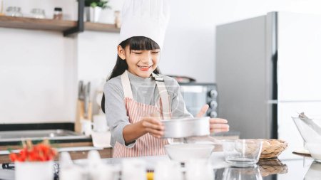 Photo for Little asian girl with chef hat and apron cooking in kitchen. Preparation for homemade bake cake. Leisure lifestyles with at home. Kid growth with interest skill concept. - Royalty Free Image