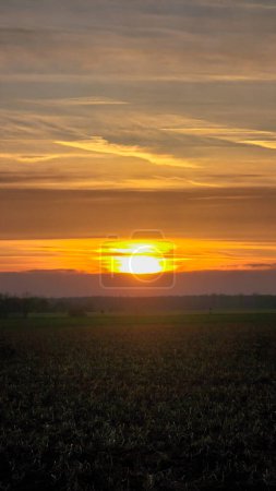 Die bernsteinfarbene Sonne geht über einer natürlichen Landschaft unter und wirft ein warmes Nachglühen auf das Feld in der Ferne, Wolken malen den Himmel in Rot- und Orangetönen Wunstorf Hannover Deutschland