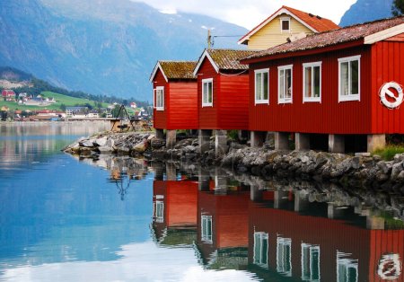 Traditional wooden red cottages on the shores of a fjord in Norway. In the background mountains and houses in a small village. Norwegian landscape