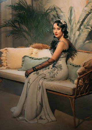 Photo for 1920s style Happy sexy flapper woman sitting on sofa, elegant gray silver dress black ostrich feather. Roar fantasy girl beauty face. classic room. white pearl beads headband, cold wave hairstyle 20s. - Royalty Free Image