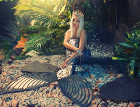 Mystical fairy woman fantasy mermaid embraced arms silver art scales fish tail. Sea goddess nymph girl sits on seabed. Blonde long flowing hair, crown. Bright sun light plants green tropical coast.