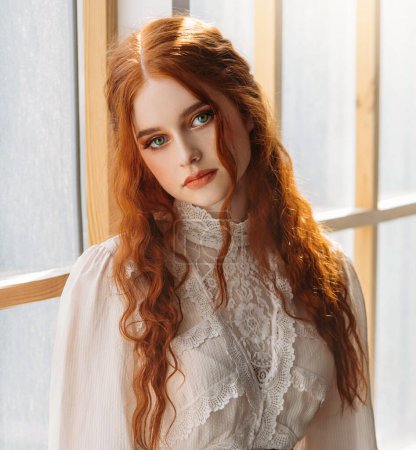 Portrait fantasy beauty red-haired woman green eyes looking at camera. White old style vintage dress. Stads by window waiting love. Curly red hair Redhead fashion model sexy girl princess beauty face