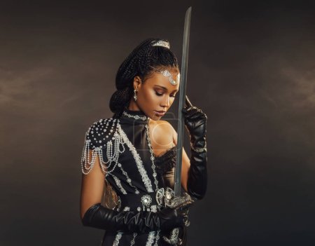 Portrait fantaisie guerrière afro-américaine tenant arme à épée dans la main. Dark queen girl en costume militaire noir. Dame gothique elfe fée. Sexy beauté visage mannequin de mode prier. Studio photo.