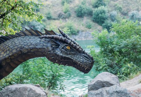 Téléchargez les photos : Fantasy art portrait real dragon head close-up sharp teeth beautiful eyes muzzle in dark gray-green scales, spikes. Creative big dinosaur toy of Jurassic period. Background summer nature green trees. - en image libre de droit