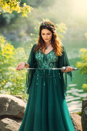 Photo for Fantasy warlike red-haired woman queen holding silver sword in hands praying before battle. Ritsa ritual of sacrifice. Green silk dress long elven sleeves, golden crown emeralds gems medieval style. - Royalty Free Image