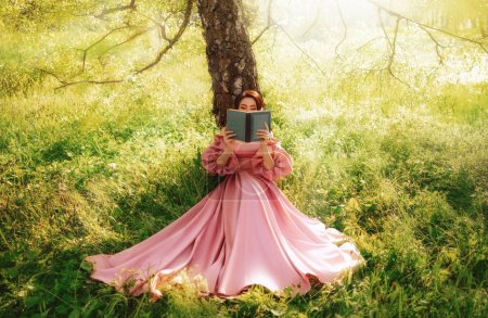 Fantasy woman sits under tree holding romantic book in hands reading novel. Pink long vintage dress. Fairy princess girl in garden summer nature green grass magic sun rays light. art photo real people