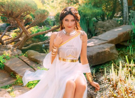 Photo for Art photo Fantasy woman greek goddess sits in spring garden green tree grass nature day magic sun light. Sexy girl queen antique style white silk dress, gold jewelry set necklace bracelet crown belt - Royalty Free Image