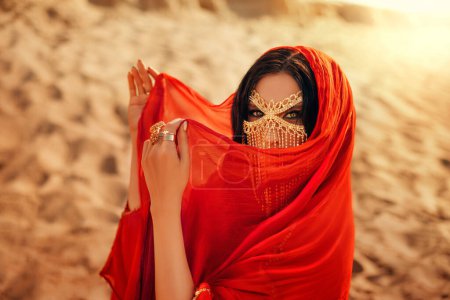 Téléchargez les photos : Mystère arabique femme beauté yeux maquillage masque doré voile niqab peau visage. Fantaisie fille art photo oriental modèle de mode cheveux noirs couverture écharpe de soie, robe rouge abaya. Dunes de sable lumière du soleil désert. - en image libre de droit