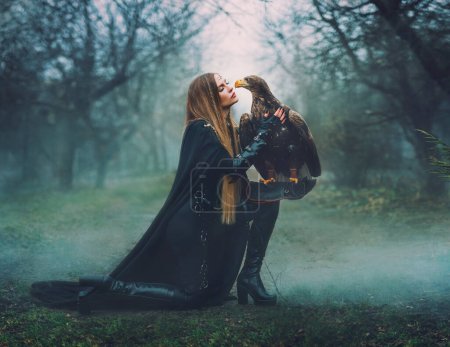 art portrait real people fantasy woman holding white-tailed eagle wild bird on hand. Elf warrior girl beauty face with brown bird pet walking in forest. queen hunter black costume dark trees fog mist