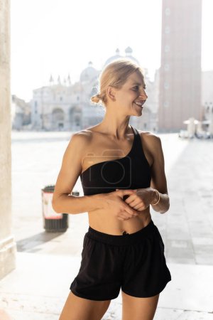 Téléchargez les photos : Femme de sport blonde positive en vêtements de sport noirs avec short noir et haut de culture regardant loin dans la rue à Venise - en image libre de droit