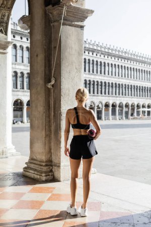Rückenansicht einer fitten Sportlerin in schwarzem bauchfreiem Top, kurzen Hosen und weißen Turnschuhen mit Fitnessmatte auf der Straße in Venedig 