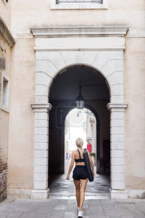 Photo for Back view of sportswoman in black crop top, shorts and white sneakers with fitness mat walking on street in Venice - Royalty Free Image